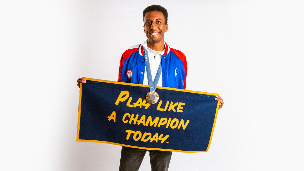 Yared Nuguse poses with his Olympic bronze medal and a Play Like a Champion Today pennant.