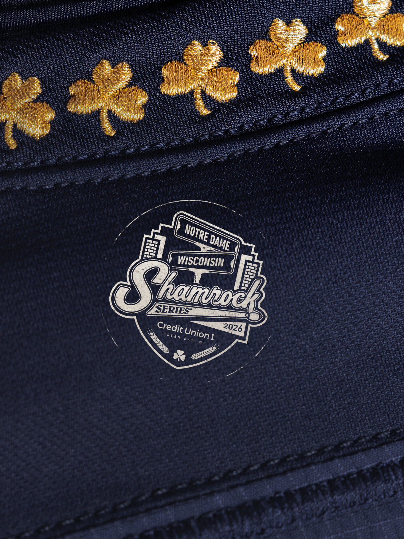 Close-up of the 2026 Notre Dame vs. Wisconsin Shamrock Series uniform patch for Credit Union 1 in Green Bay, Wisconsin, shown on navy fabric beneath gold embroidered shamrocks.